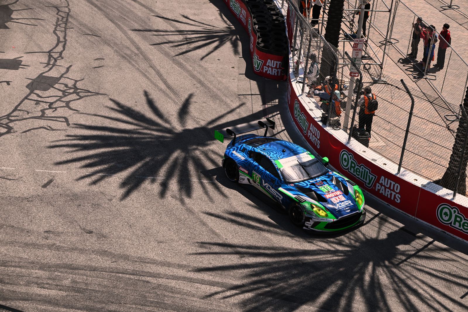 The #19 Aston Martin from above at Long Beach, crossing palm tree shadows beside the wall